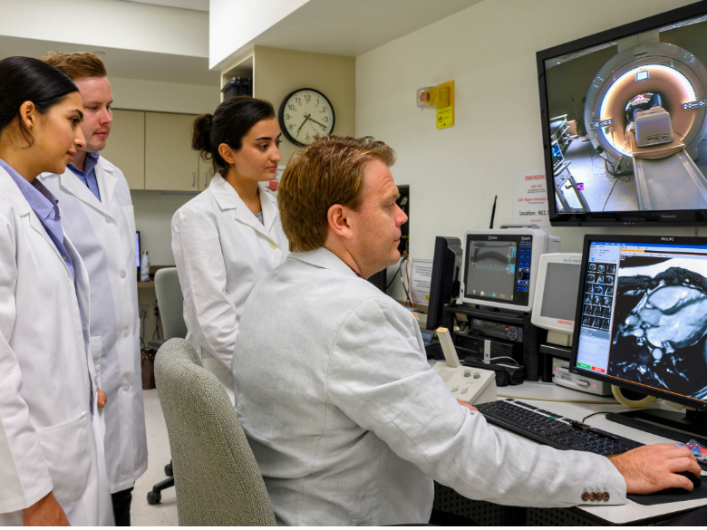 A group of individuals in lab coats observing a medical imaging workstation displaying MRI scans.