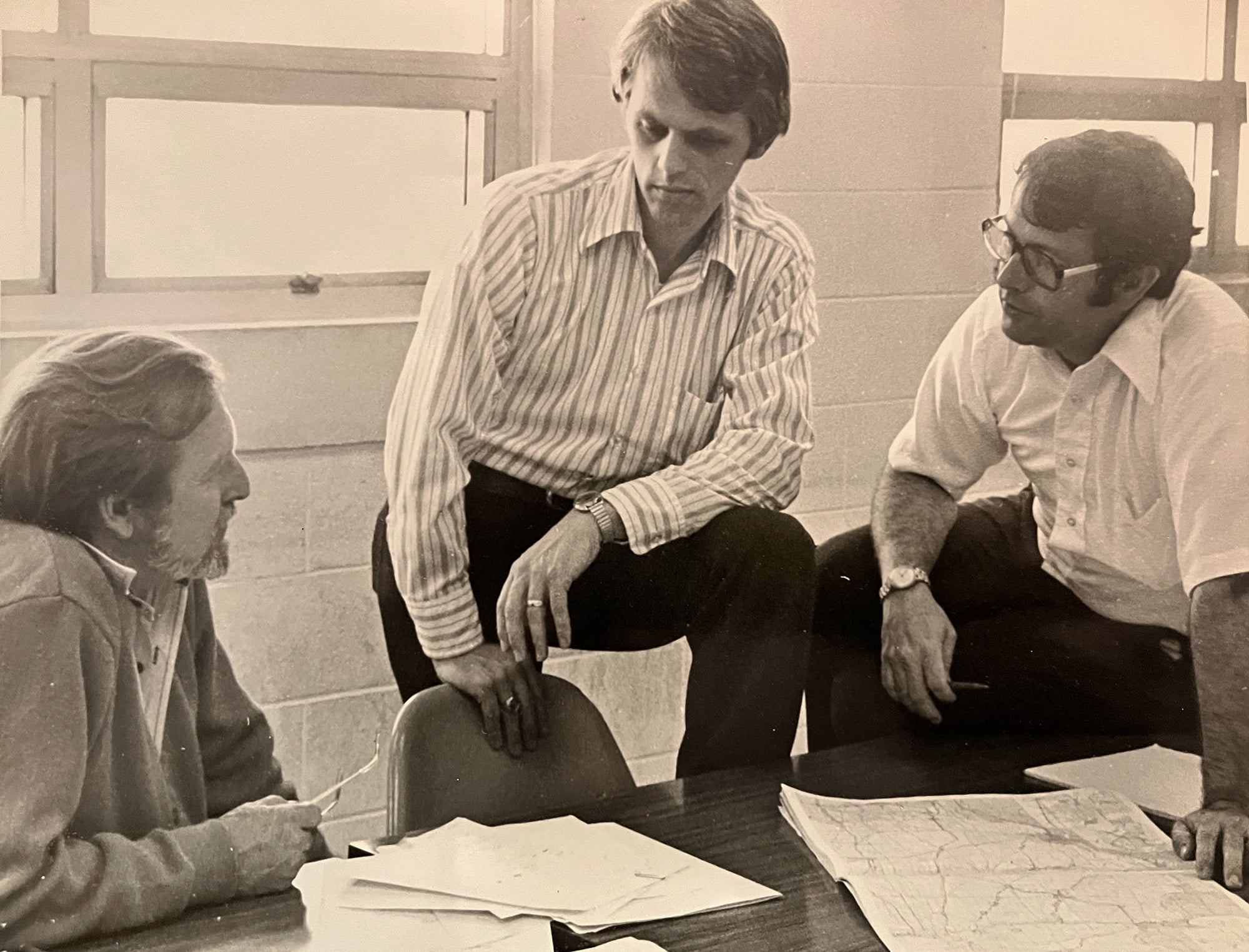 In a black and white archival photo, researchers Garvin McCain (left), Paul Paulus (center), and Verne Cox (right) are clustered around an office desk, intently discussing papers and a map laid out before them.