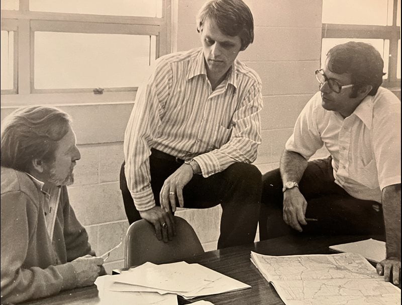 In a black and white archival photo, researchers Garvin McCain (left), Paul Paulus (center), and Verne Cox (right) are clustered around an office desk, intently discussing papers and a map laid out before them.