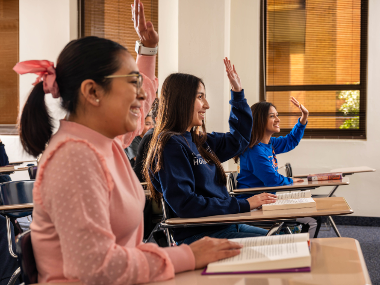Students seated at desks in a classroom raise their hands while reading from open books.