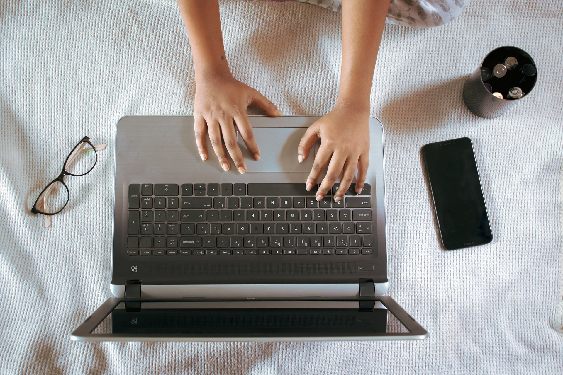 person typing on a laptop with glasses, cup, and cellphone next to it