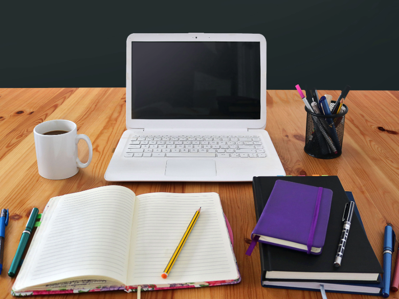 Open notebook with a yellow pencil, laptop, coffee mug, pens, and stacked notebooks arranged on a wooden desk against a dark background.