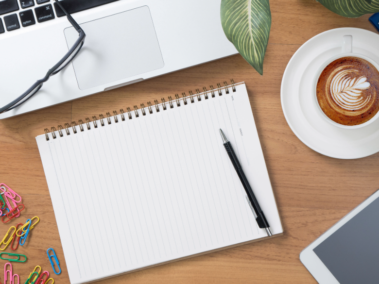 Top-down view of a workspace with a blank spiral notebook and black pen, surrounded by a laptop, coffee cup, green plant, colorful paper clips, and part of a tablet.