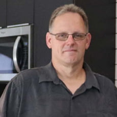 picture of Brad McCorkle, dressed in a dark button up in front of cabinets. He stands in a house his students helped build for a service learning project.