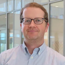 picture of Justyn Jaworski, dressed in a formal button up and dark-rimmed glasses in front of tall windows