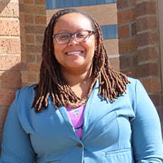 picture of Kyrah Brown, dressed in a blazer that's buttoned over a floral blouse with glasses in front of brick pillars