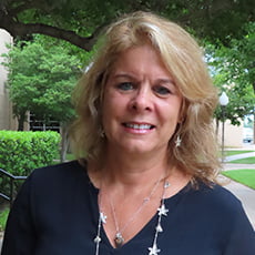 picture of Pamela Johnson, dressed in a dark formal shirt with a beaded necklace and earrings in front of trees and bushes