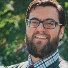 picture of Timothy Ponce, dressed in a vest, plaid shirt, and tie. He ahs glasses and stands in front of trees