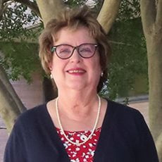 picture of Joyce Myers, dressed in a cardigan over a floral shirt with a pearl necklace, earrings, and glasses