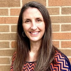 picture of Alice Lubbe, dressed in a zig-zagged shirt with earrings on in front of a brick wall