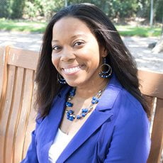 picture of Brandie Green, dressed in a blazer with matching earrings and necklace, sitting on a bench on campus