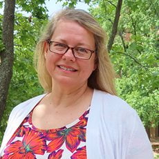 picture of LaDonna Aiken, dressed in a cardigan over a floral shirt with dark-rimmed glasses in front of trees on campus