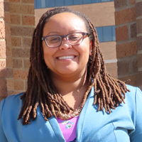 picture of Kyrah Brown, dressed in a blazer that's buttoned over a floral blouse with glasses in front of brick pillars