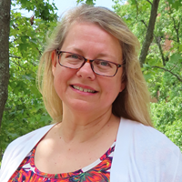 picture of LaDonna Aiken, dressed in a cardigan over a floral shirt with dark-rimmed glasses in front of trees on campus