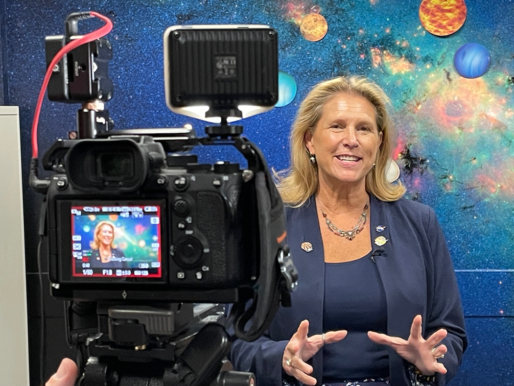 Dr. Lori Glaze presenting on camera in front of solar system art.