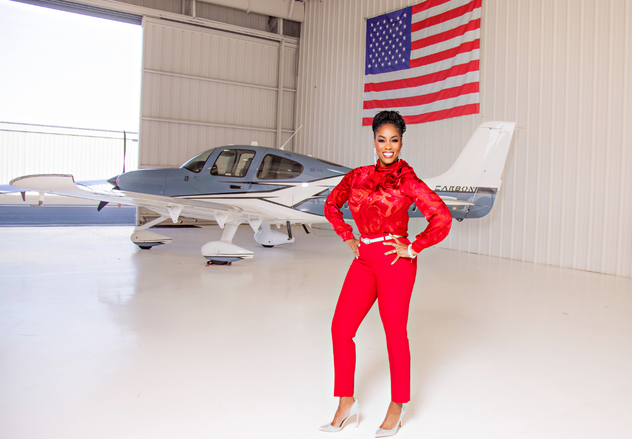 Tamiko Bailey standing in front of a plane in a red suit. Entire plane is in frame in a warehouse with American flag hanging