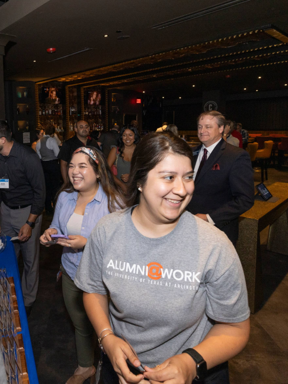 Alumna smiling and looking to her left in a crowded room. She is wearing an Alumni@Work shirt.