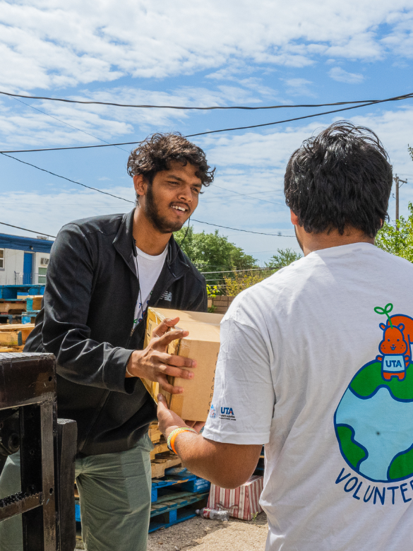 Two volunteers passing a box between each other on a bright, sunny day outside. One is facing the camera and smiling at the other person.