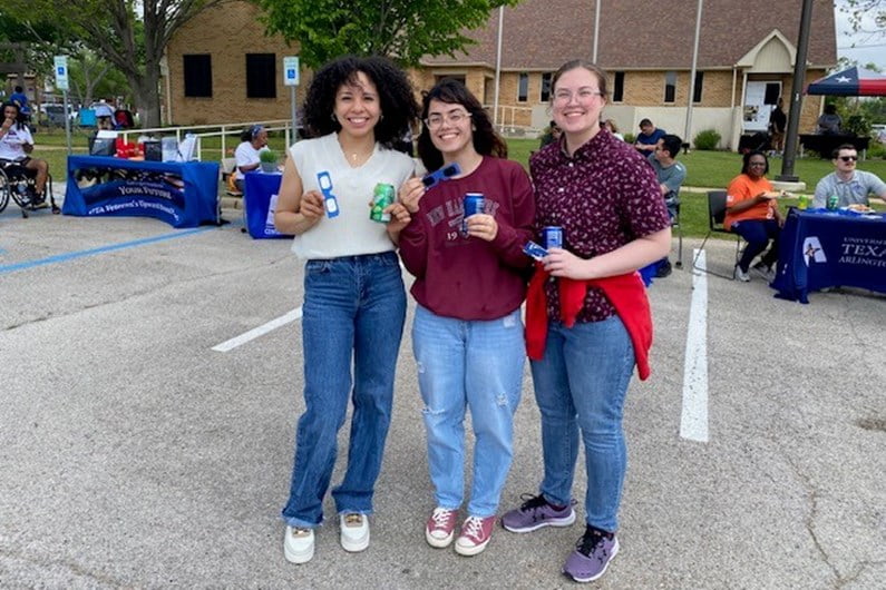 Three women standing closely together smile at an outdoor event. They hold canned drinks, with tables and people seated in the background. It's a lively and cheerful scene.