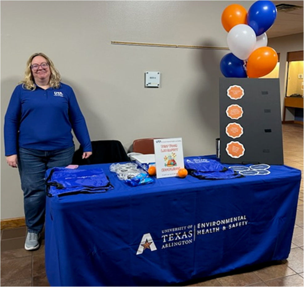 A woman stands smiling beside a table with a University of Texas Arlington Environmental Health & Safety tablecloth. The table holds balloons, informational materials, and giveaways.