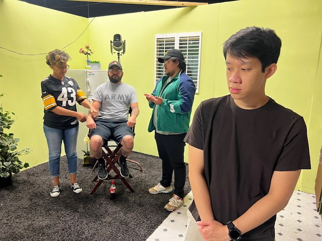 Inside a small green-walled set, an actor sits in a director’s chair as two crew members talk and another student stands off to the side.