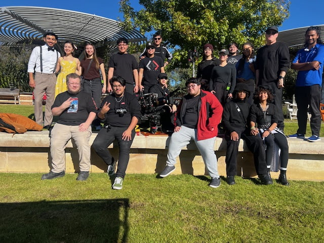 A group of film students standing and sitting outdoors around a professional camera rig under a pergola on a sunny day.