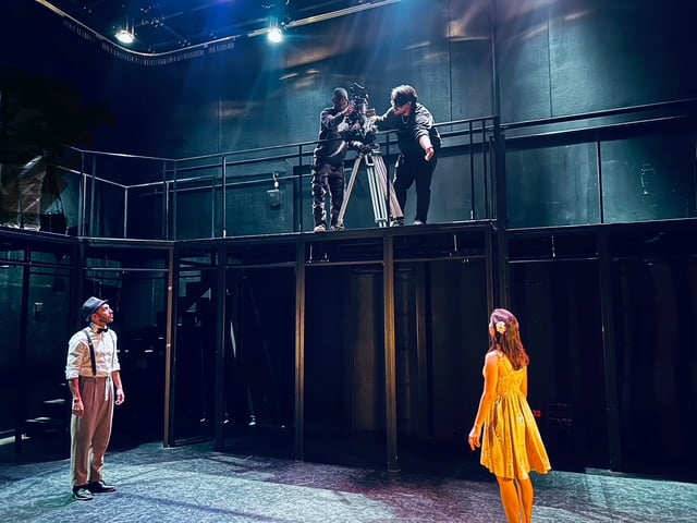 On a darkened stage, two actors one in vintage attire and one in a yellow dress perform while crew members above adjust a camera on a catwalk.