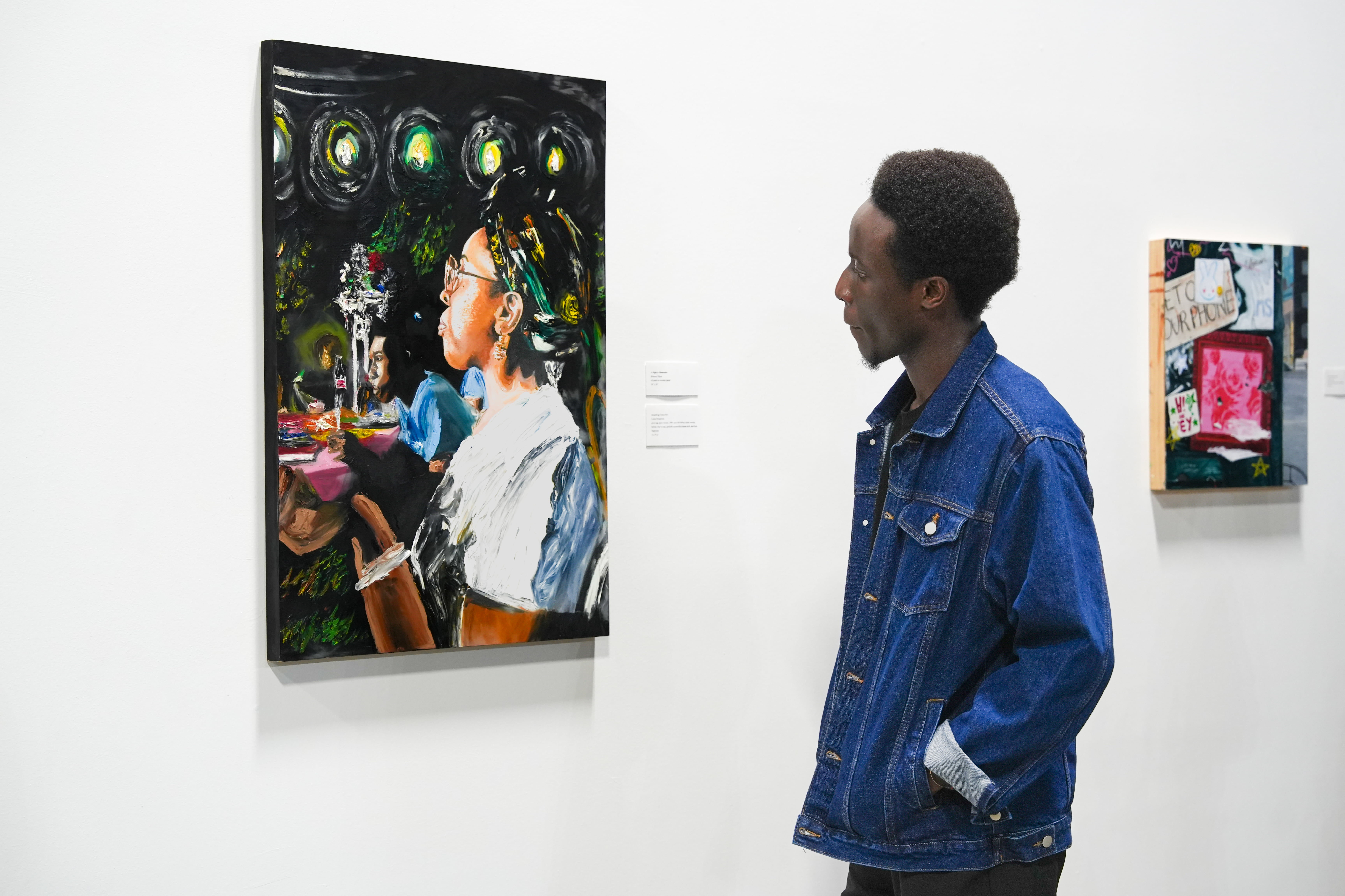 A student in a denim jacket looks closely at a vibrant oil painting of a woman sitting at a table under bright lights.