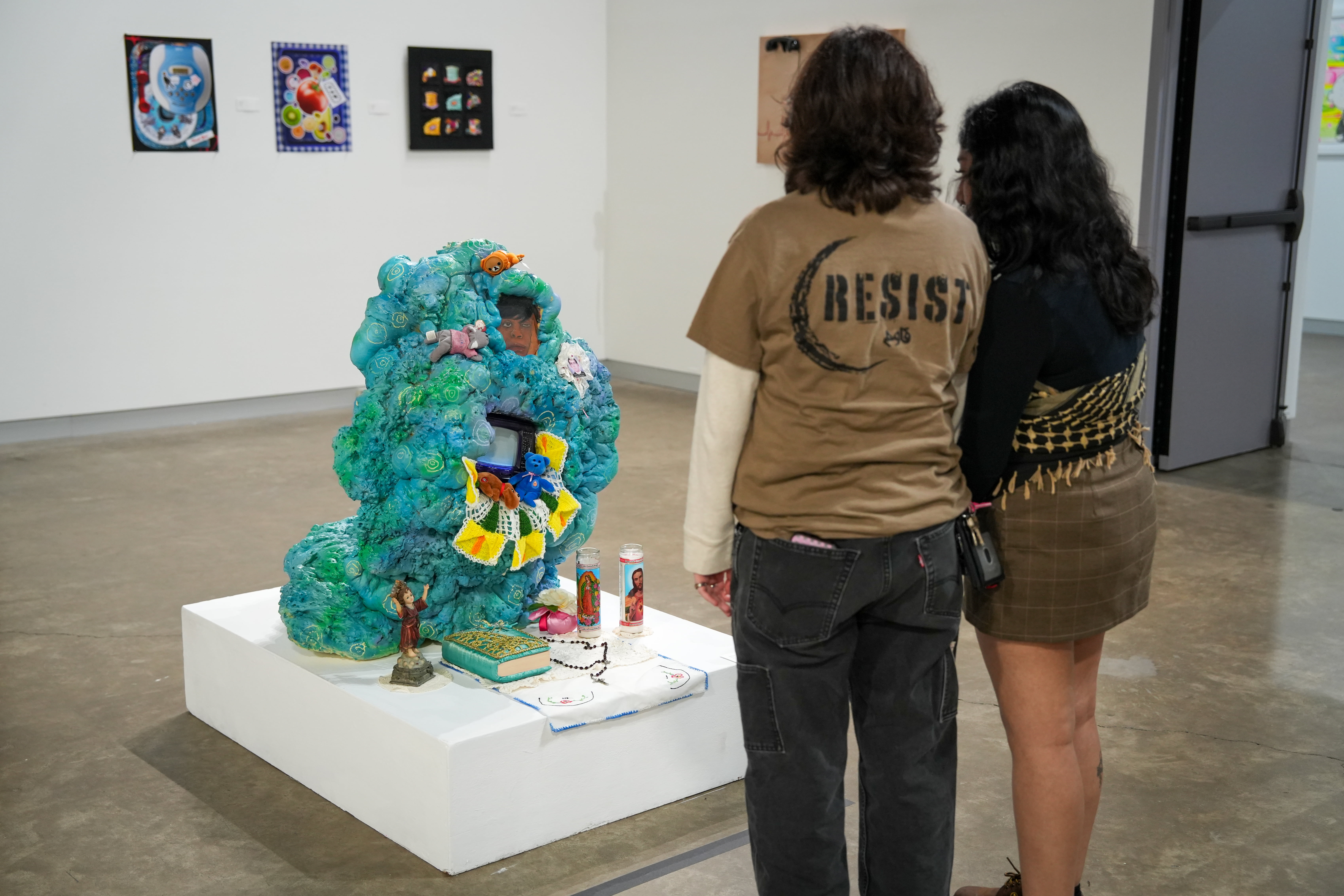 Two visitors stand before a blue, textured multimedia sculpture adorned with small photos, toys, and religious candles.