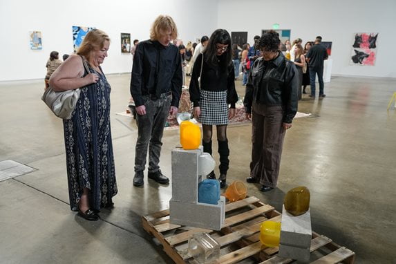 Four visitors gather around a floor installation featuring colorful blown glass shapes resting on concrete blocks and a wooden pallet.