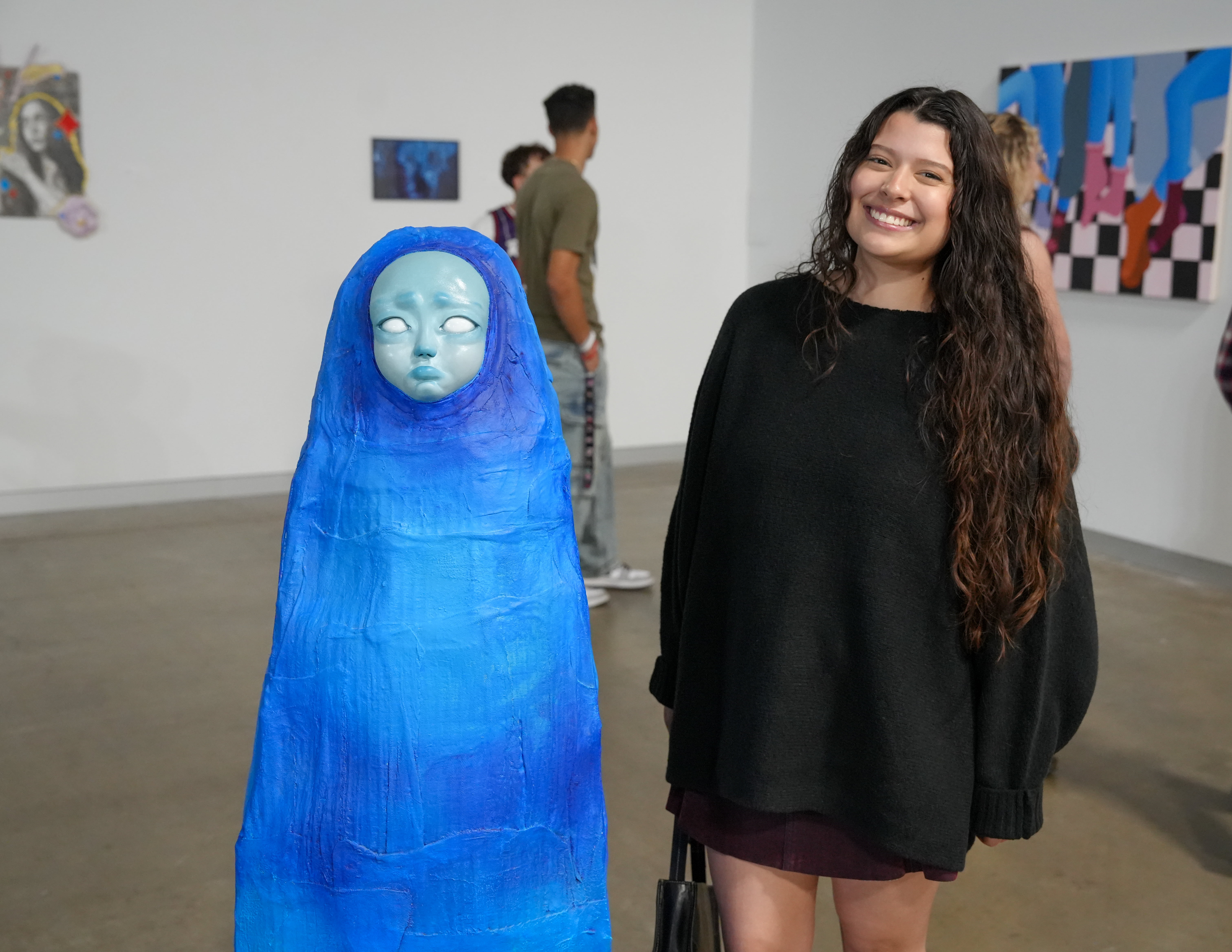 A smiling student stands next to a tall, bright blue sculpture of a shrouded figure with a pale, melancholic face.