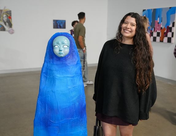 A smiling student stands next to a tall, bright blue sculpture of a shrouded figure with a pale, melancholic face.