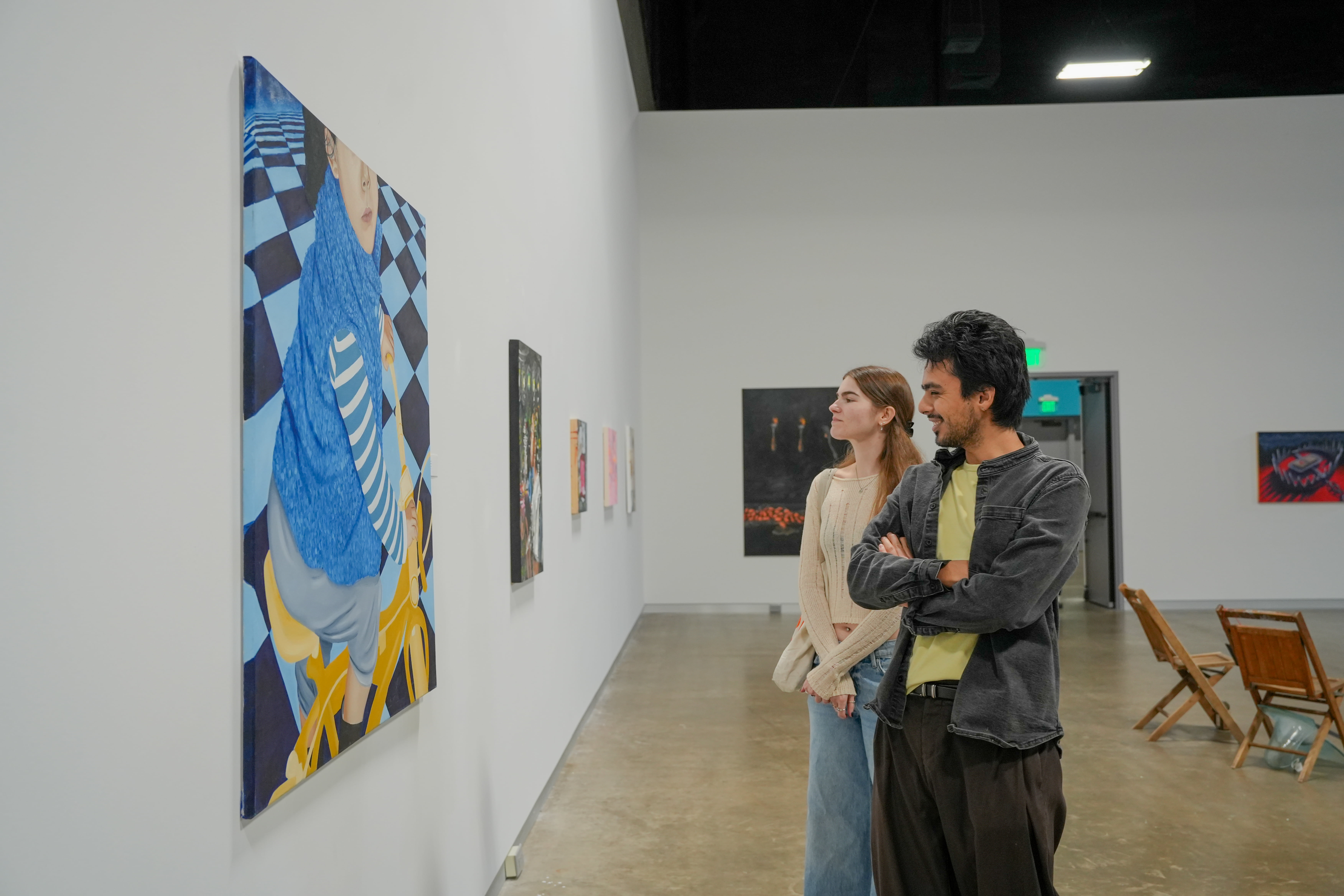 Two students admire a large painting of a figure on a yellow bicycle against a blue and black checkered background.