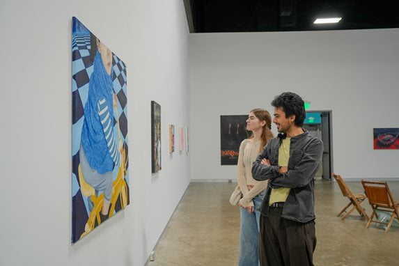 Two students admire a large painting of a figure on a yellow bicycle against a blue and black checkered background.