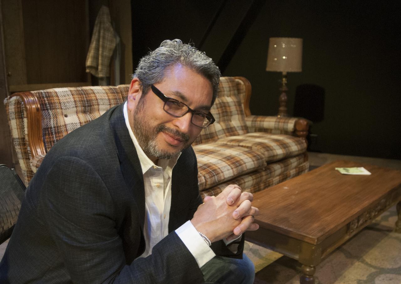 Octavio Solis wearing glasses and holding his hands together, looking at the camera with a smile