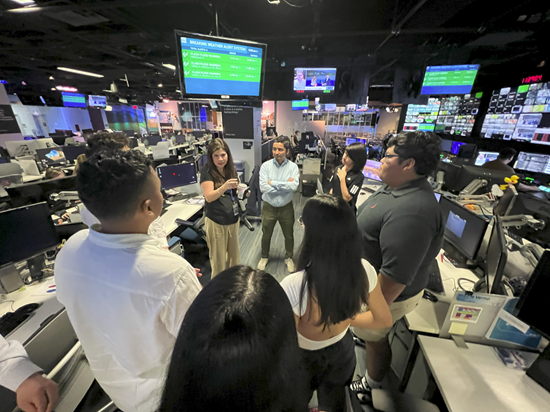 Students in a circle in a news room with lots of screens, listening to a woman speak.