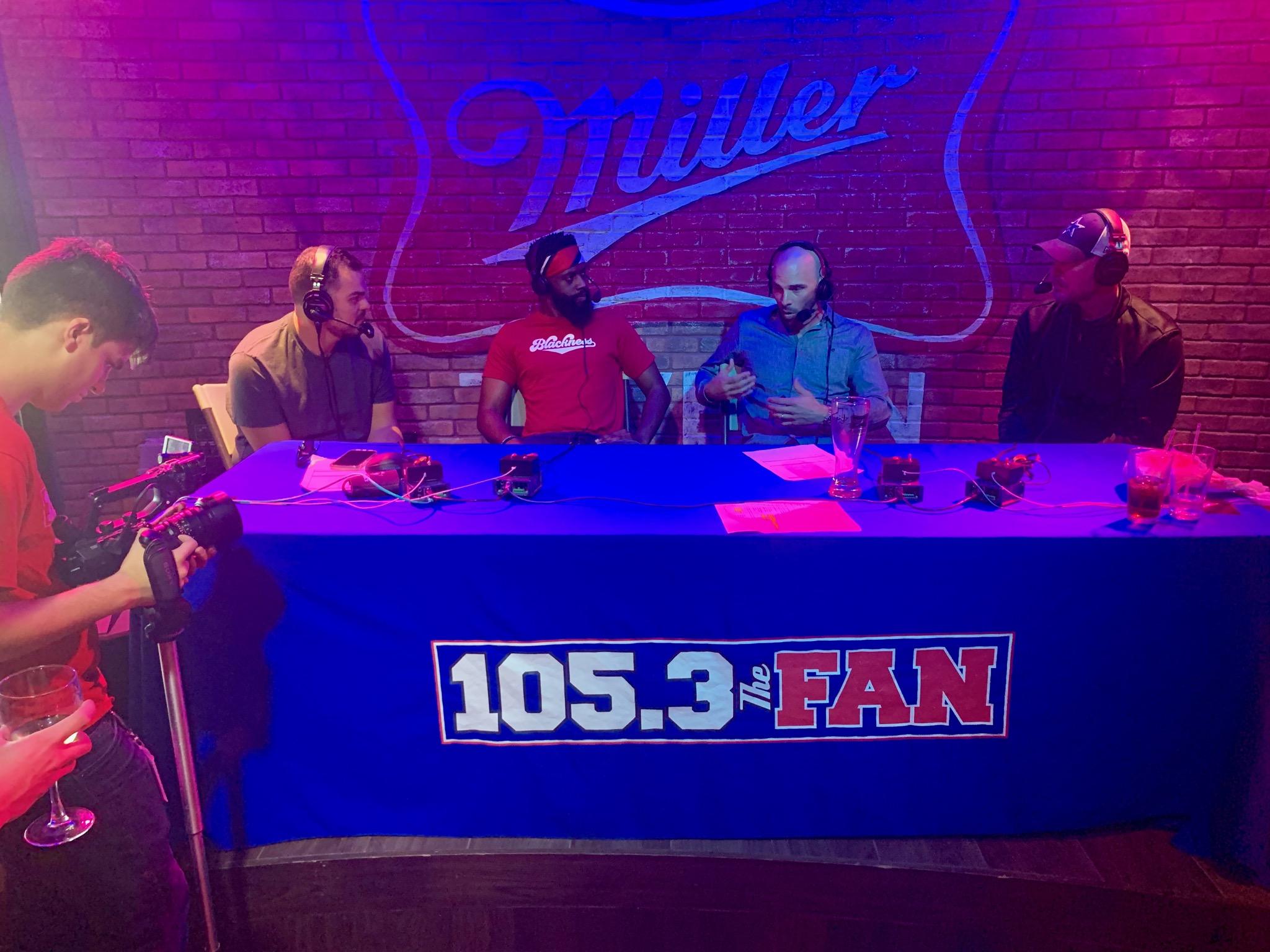 Reginald Adetula and others sat around a table with headphones on while a person films them. The tablecloth reads, "105.3 The Fan." 