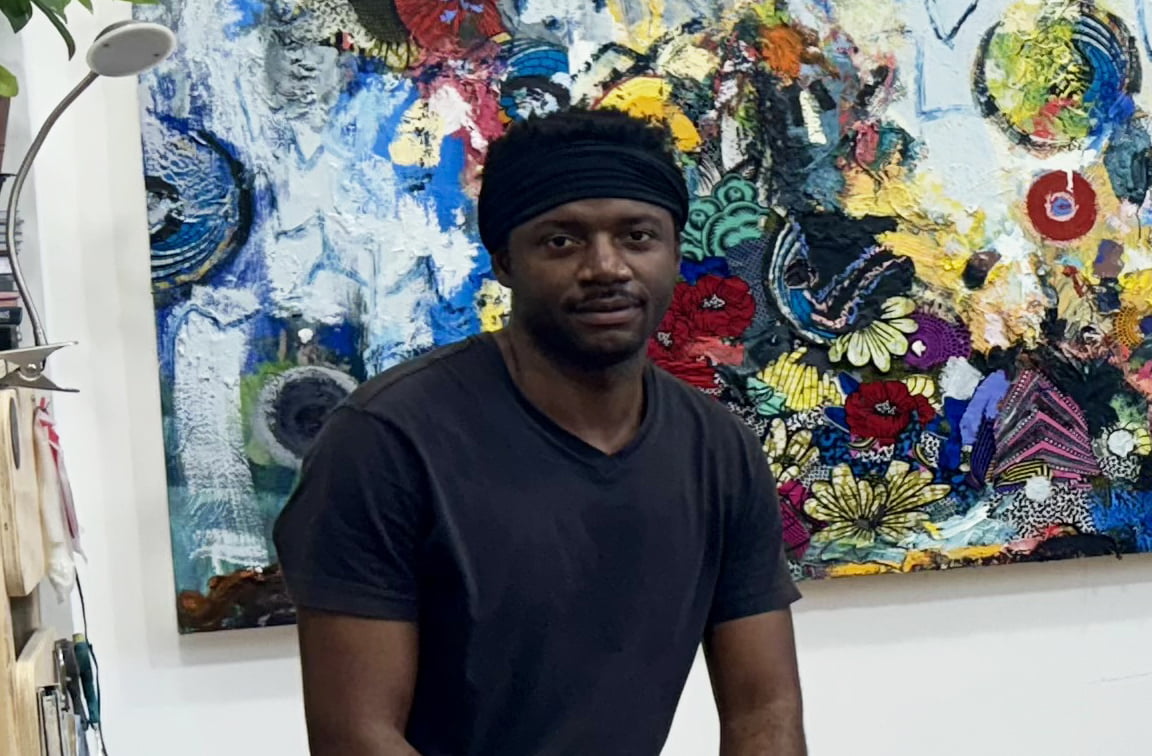 Jimi Kabela sitting on a stool in an art studio, surrounded by various paintings and painting supplies.