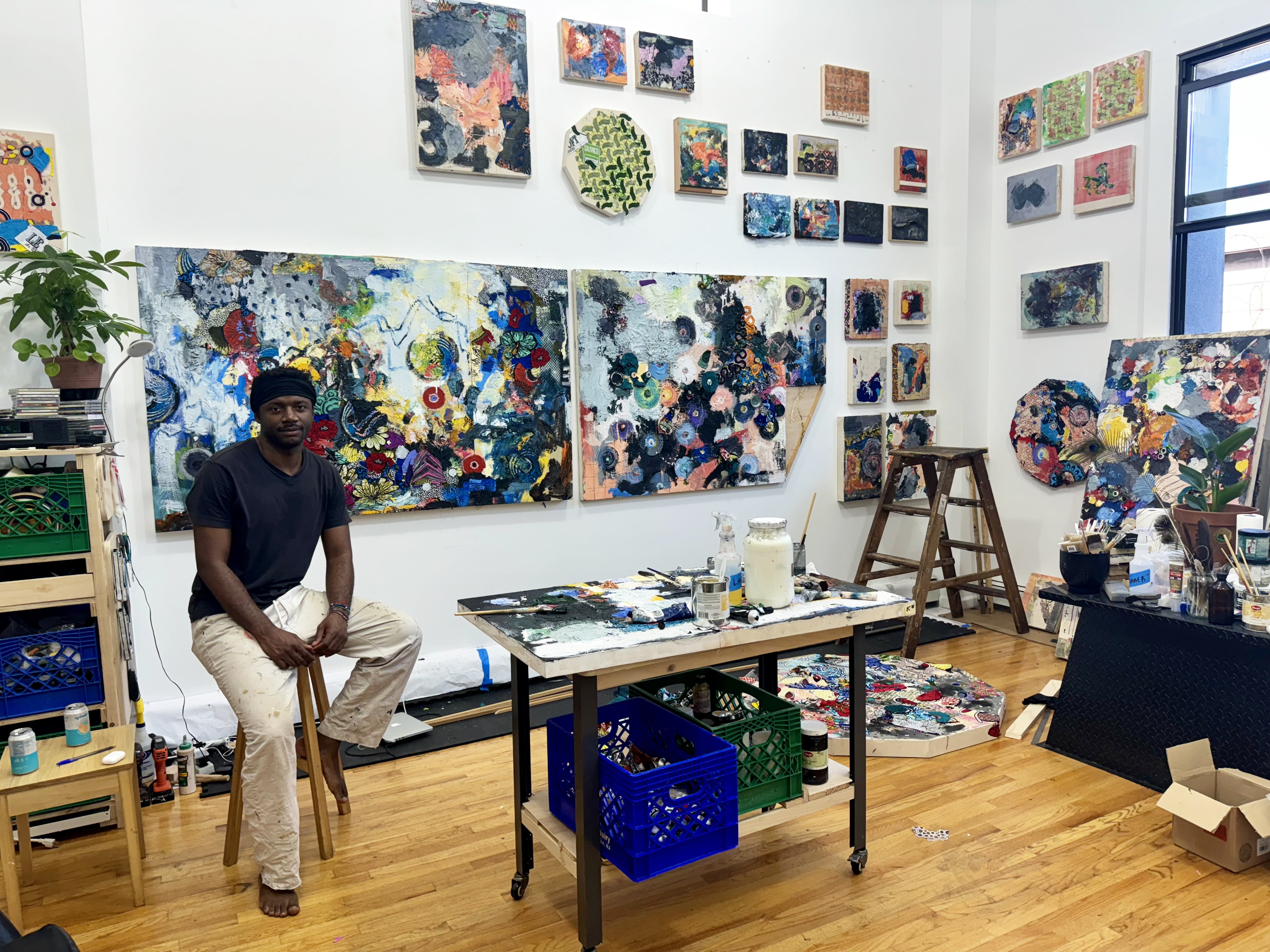 Jimi Kabela sitting on a stool in an art studio, surrounded by various paintings and painting supplies.