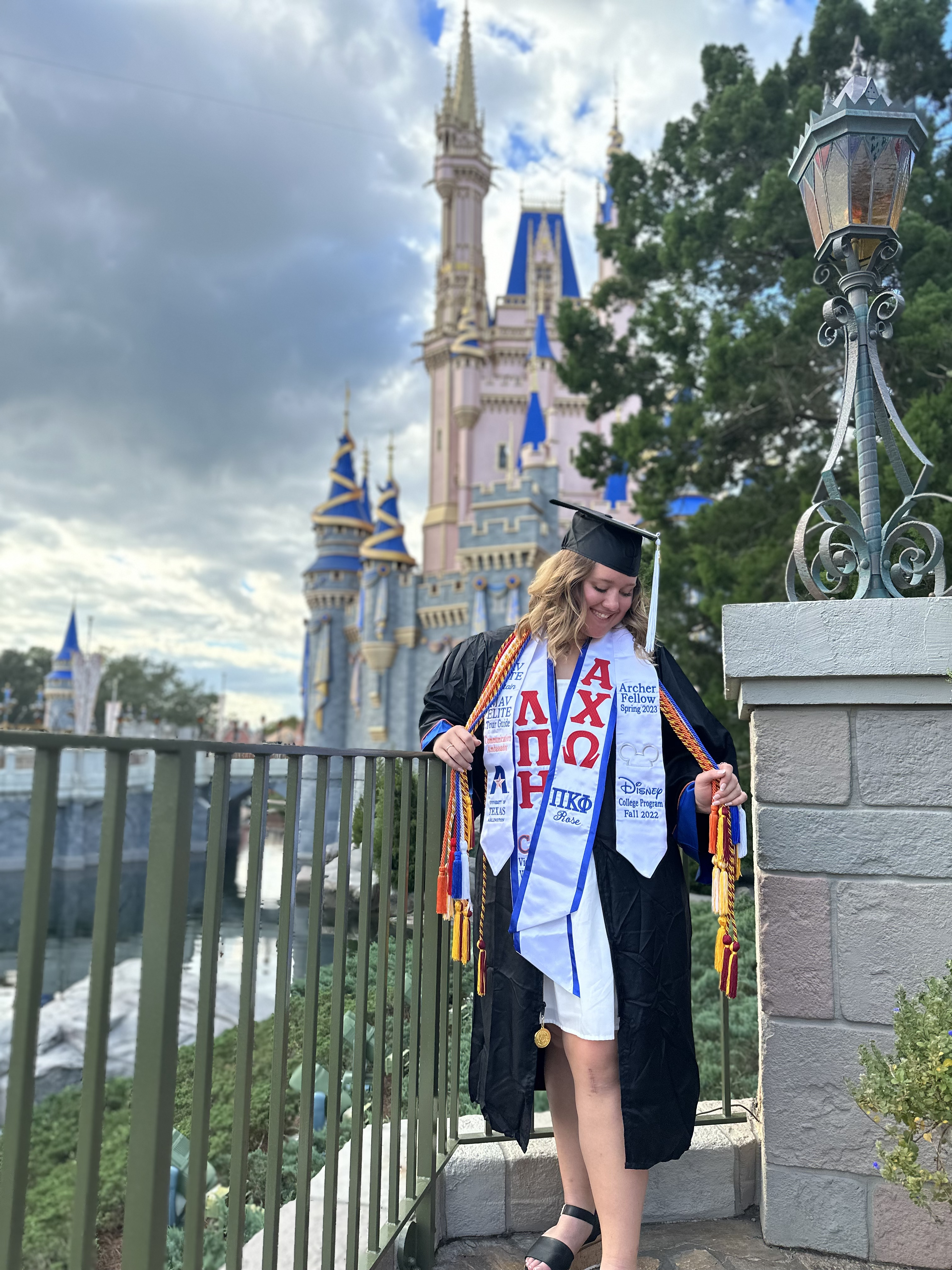 Madison Simms dressed in her cap and gown in front of a Disney castle. She wears several academic cords, a sorority stole, and a Disney Archer fellowship stole.