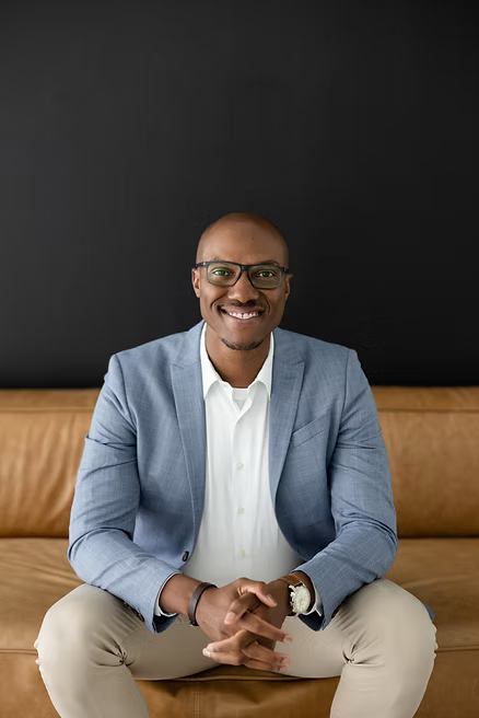 Nathan Hervey sitting on a couch, hands clasped together, smiling.
