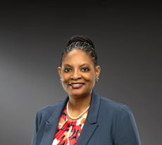 Angela Hamilton wearing a patterned blouse, blazer, and a pearl necklace smiles at the camera.