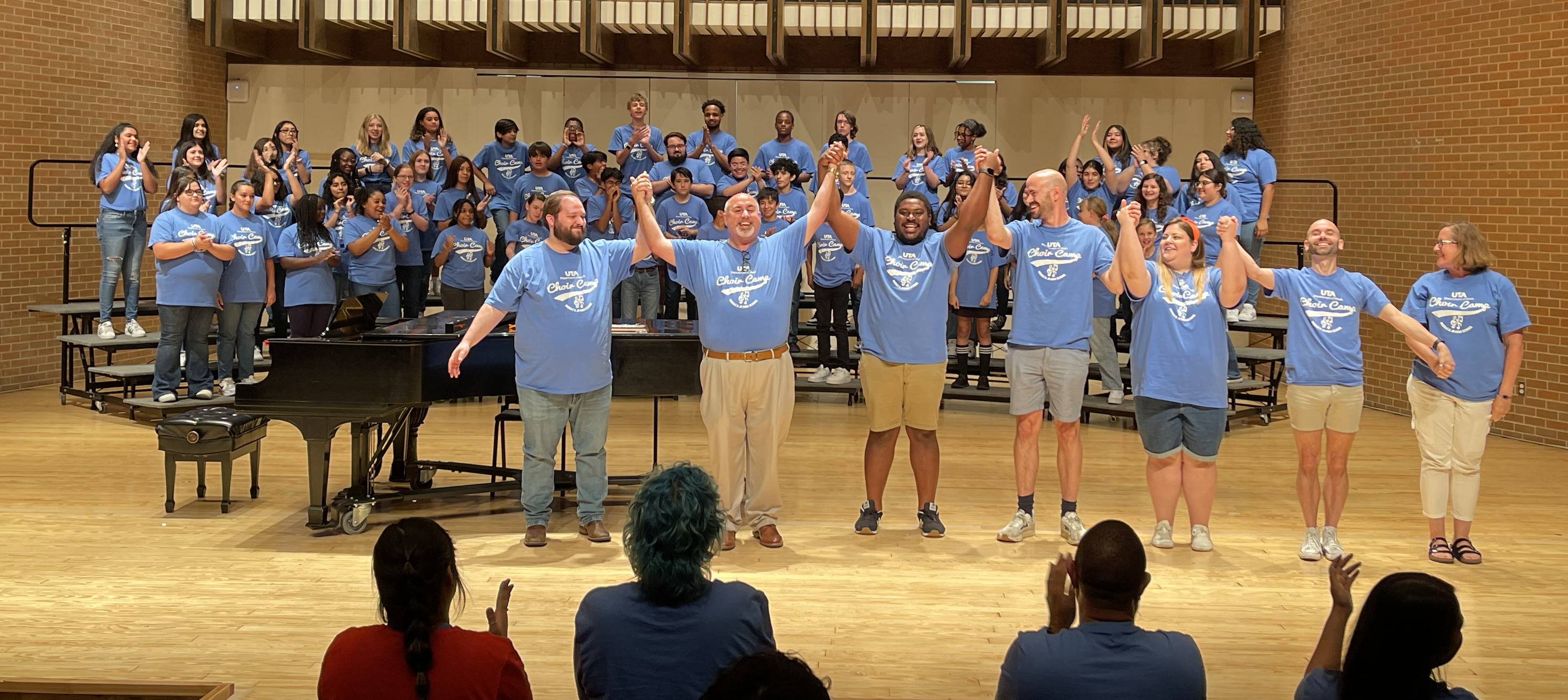 Music professors bowing on stage with students behind them, audience is giving a standing ovation.