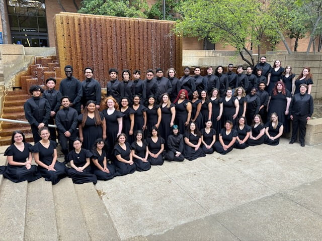 Group of students all wearing the same color, black, posed outside the water foundation by the Fine Arts building.