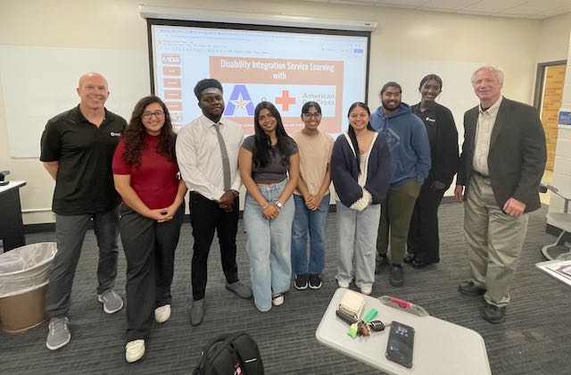 Nine people standing and smiling in front of a projector screen that has a powerpoint that's titled "Disability Integration Service Learning with U T A and American Red Cross"