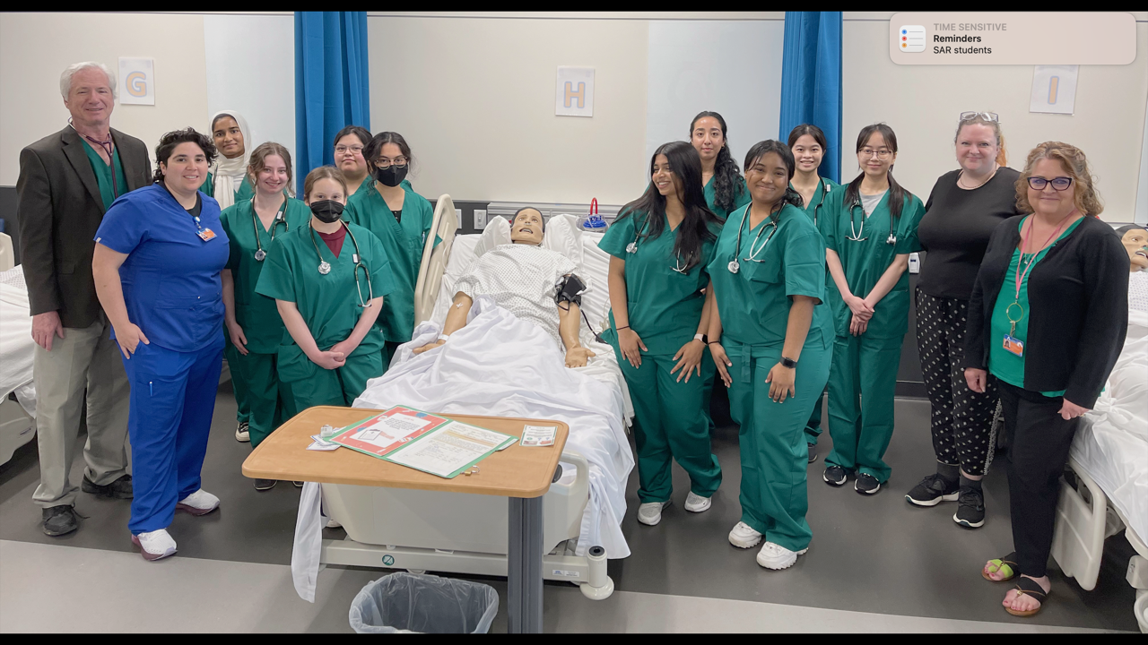 A group of students and teachers standing around a fake patient in a hospital bed.