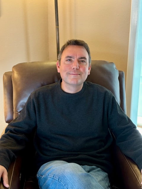 Jeremy Byrd sitting in a chair smiling while sitting in a chair with short hair, a sweater, and jeans.