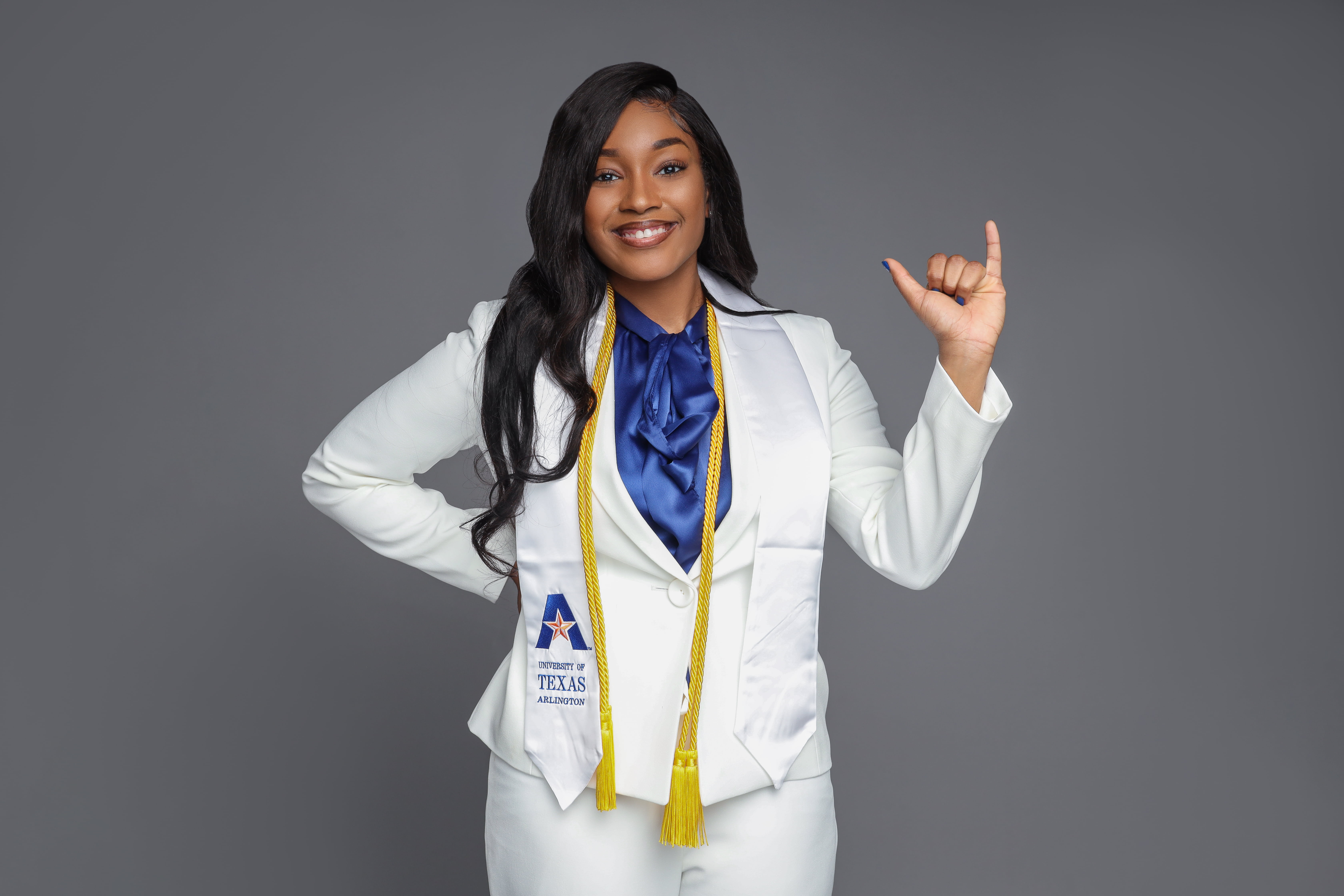 Serita Johnson smiling as she "Mavs Up," dressed in a suit, U T A stole, and two academic honor cords