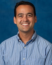 Steven Arxer in a dress shirt with short hair, smiling in front of a plain background