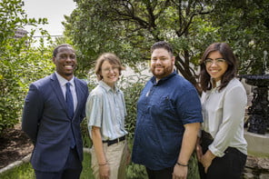 Four people stand and smile at the camera with trees in the background